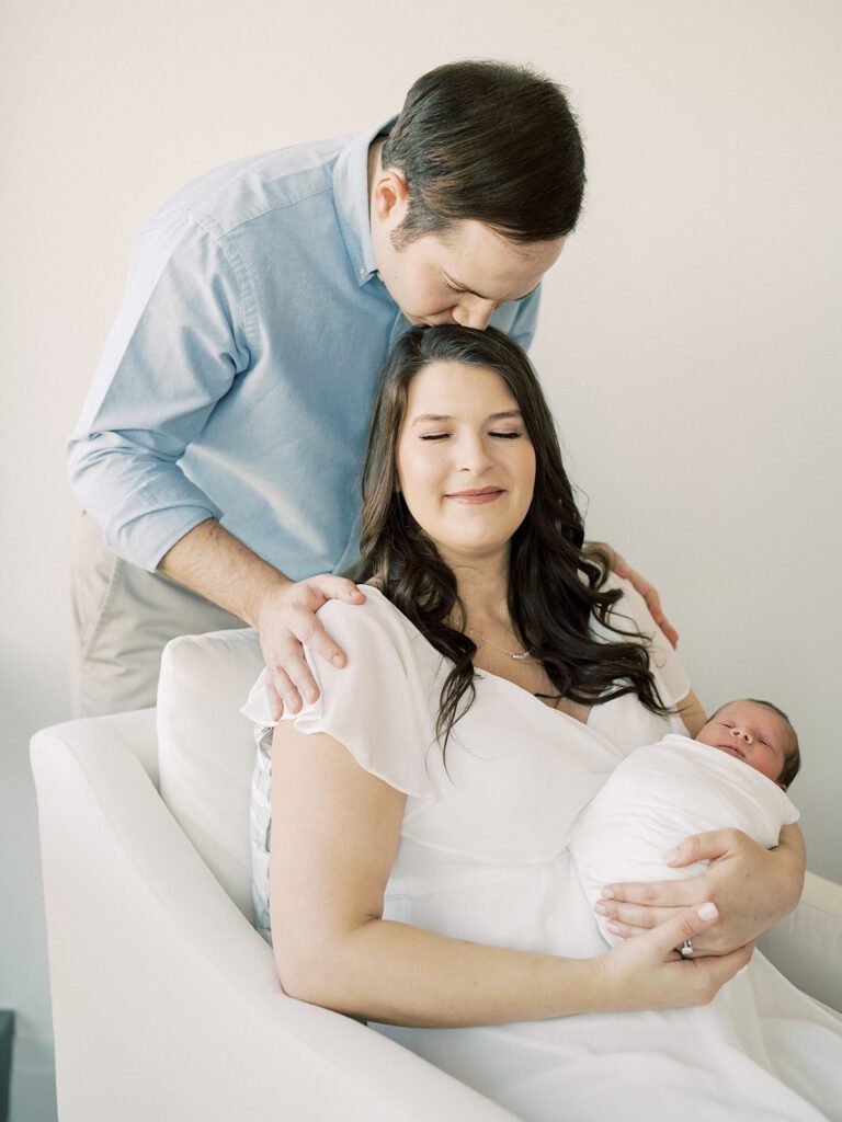 Father Leans Down To Kiss The Top Of His Wife's Head As She Sits In A Chair And Holds Their Newborn Baby During Their Georgetown Newborn Session.