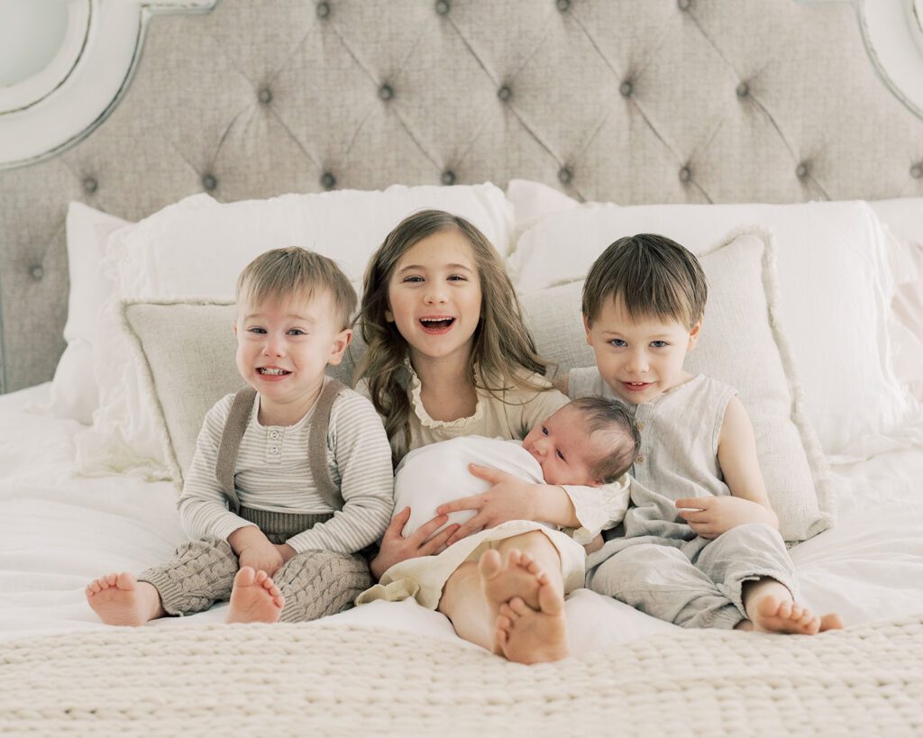 Big Sister And Two Big Brothers Sit On Bed Holding Newborn Baby.