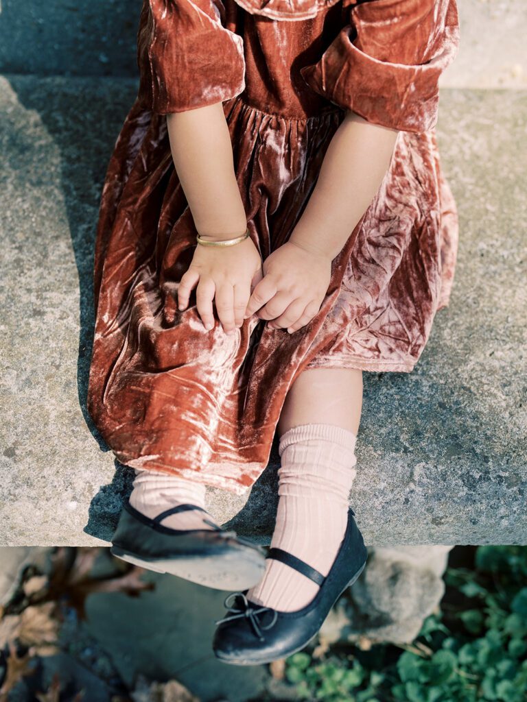 Close-up view of little girl in maroon velvet dress sitting on a stone balcony.