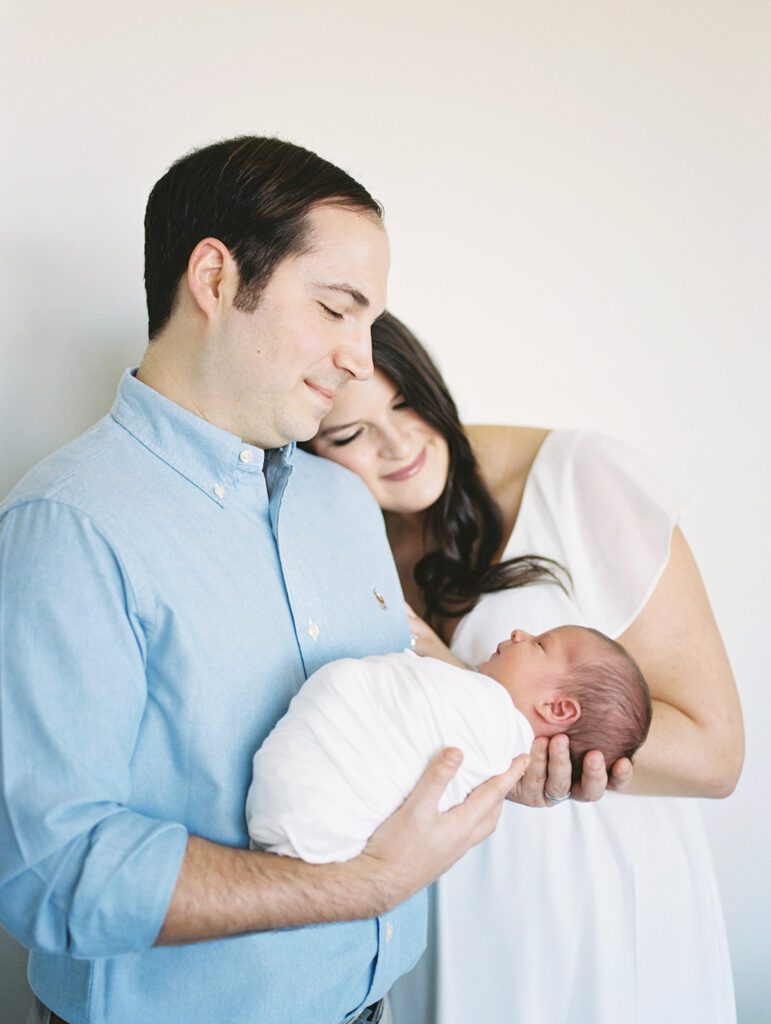 Father Smiles Down At Newborn Baby Swaddled In White As His Wife Leans Against Him During Their Georgetown Newborn Session.