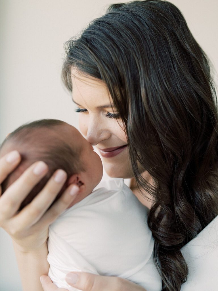 Mother With Long Brown Hair Brings Her Swaddled Newborn Son Up To Her Nose During Their Georgetown Newborn Session.