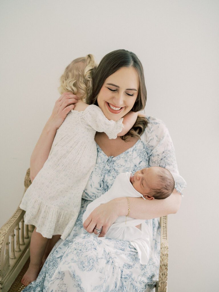 A Brown-Haired Mother Smiles Down At Her Newborn Son As Her Toddler Daughter Gives Her A Hug.