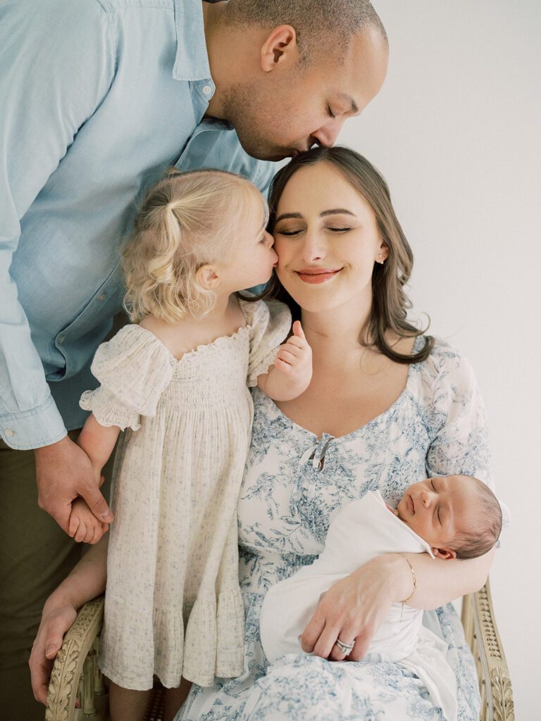 A Father Kisses The Top Of A Mother's Head As Their Daughter Kisses Her Cheek While She Holds Her Newborn Son During Their Newborn Photos In Arlington.