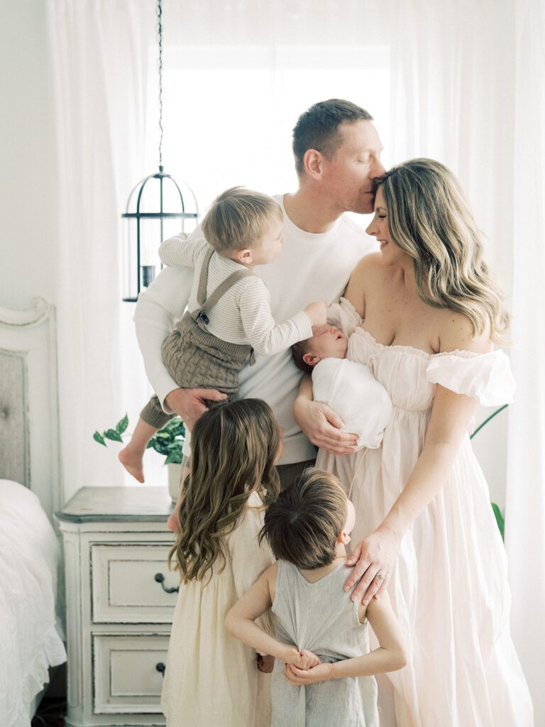 Father Kisses Wife On Forehead As He Holds Young Son And She Holds Newborn Baby While Their Two Other Children Stand Close During Their Newborn Photos In Annapolis.