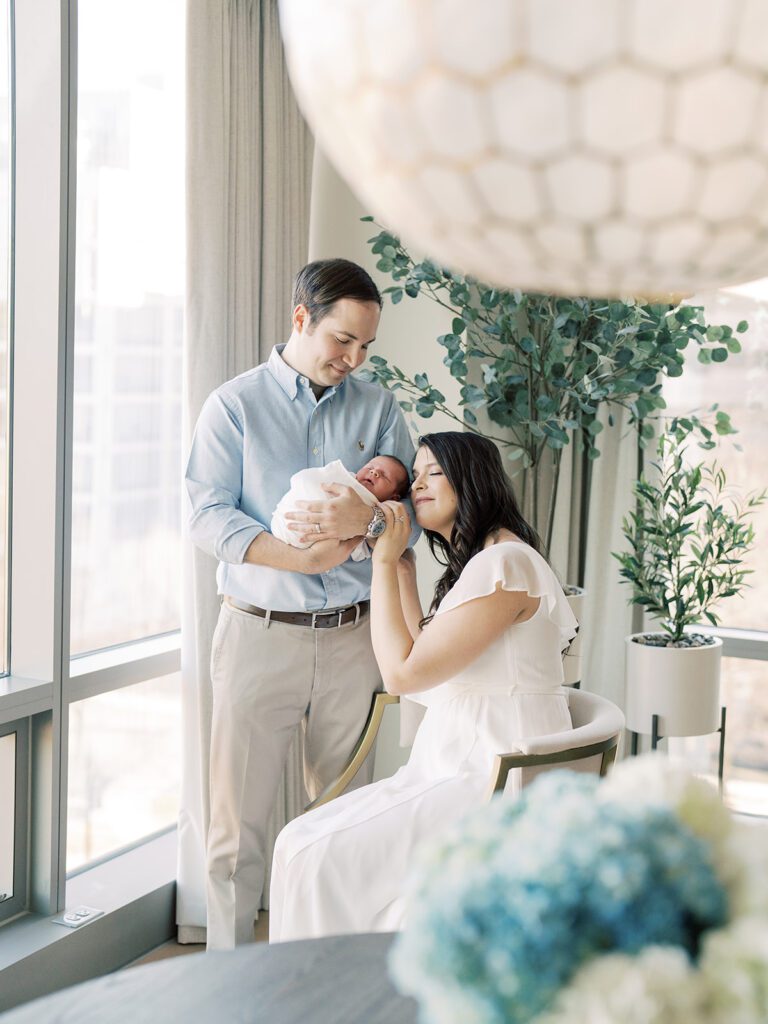 Mother Sits In A Chair And Leans Against Her Husband Who Stands And Holds Their Newborn Baby In Their Home During Their Georgetown Newborn Session.