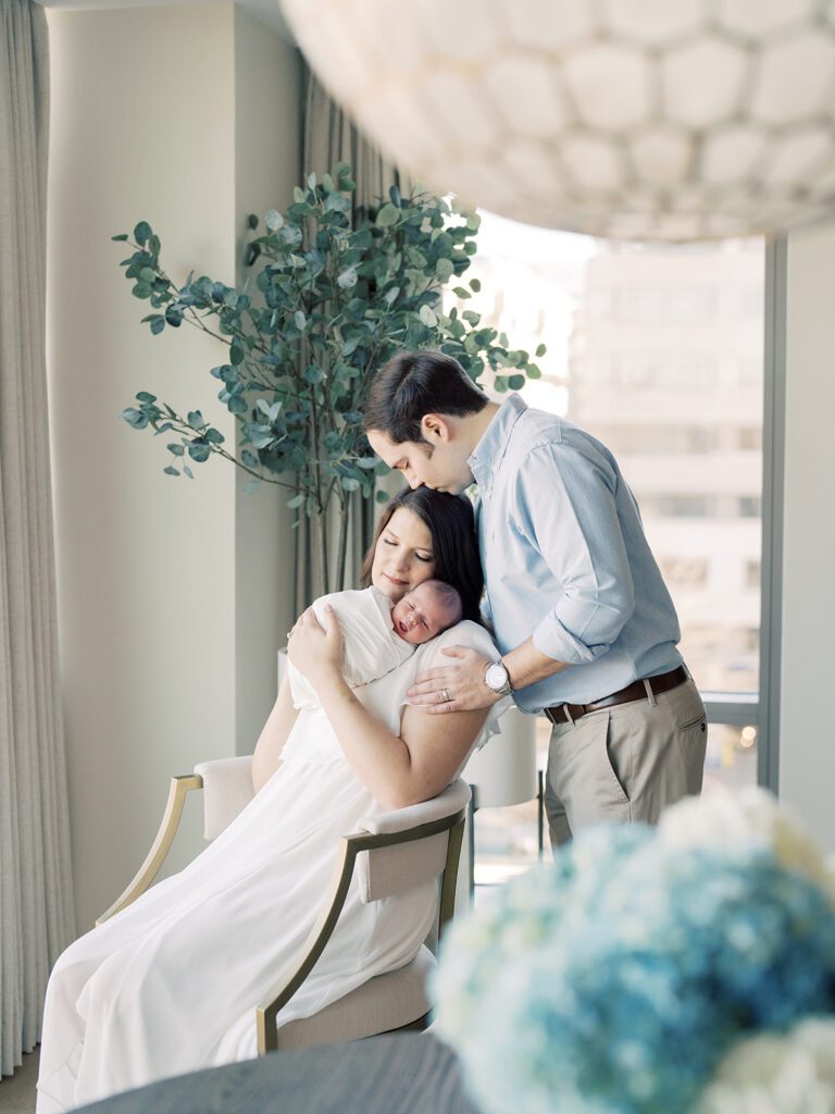 Father Kisses His Wife's Head As She Sits In A Chair In Their Home, Holding Their Newborn Baby During Their Georgetown Newborn Session.