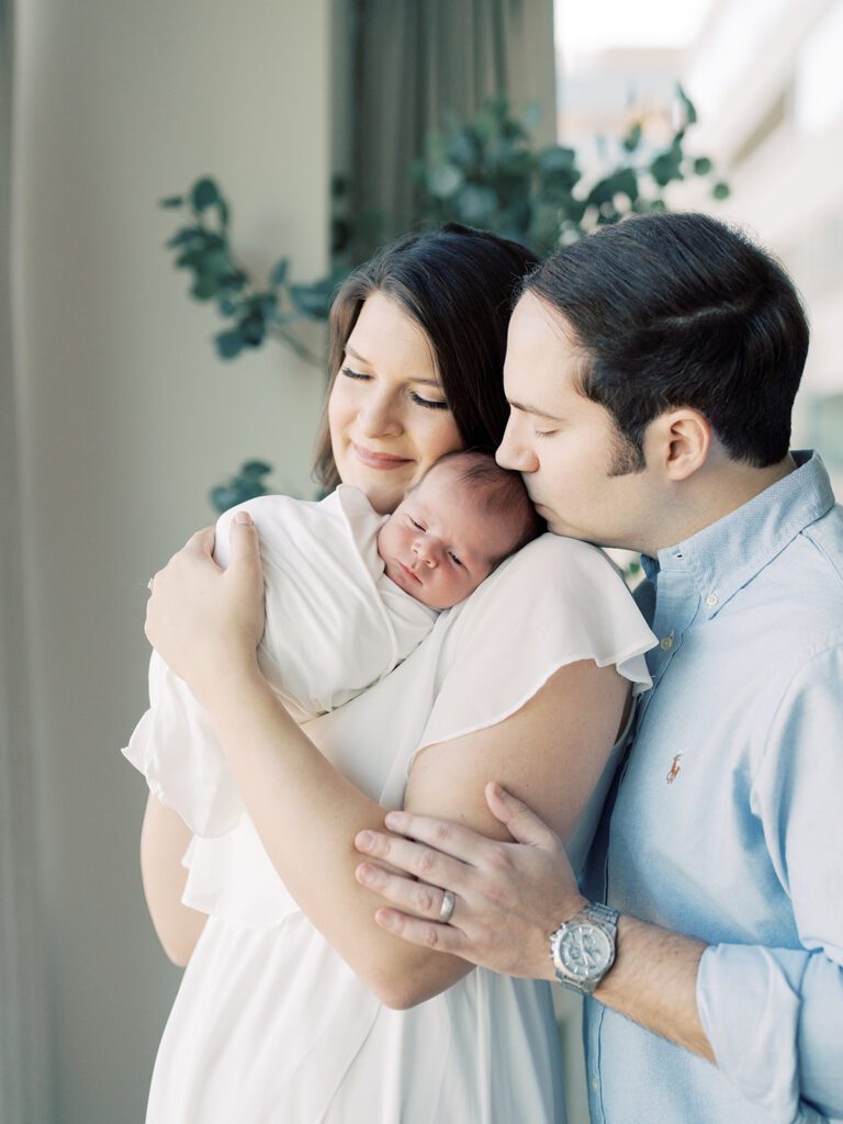 Father Stands Beside His Wife And Kisses Their Newborn Baby's Head As Mother Holds Baby In Her Arms During Their Georgetown Newborn Session.