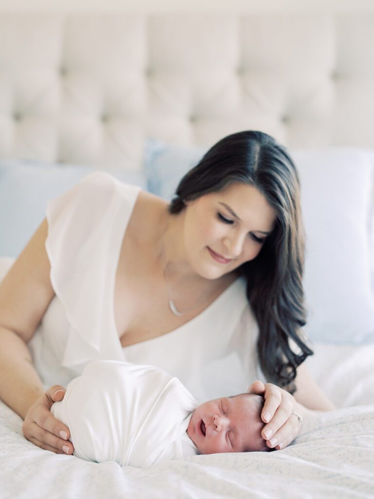 Mother With Long Brown Hair In A White Dress Places A Hand On Her Son's Head As They Lay On A Bed Together.