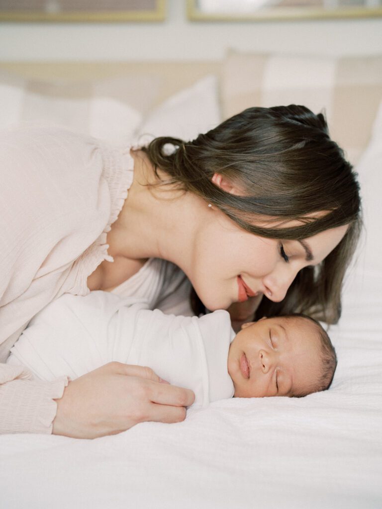 A Mother With Brown Hair Leans Down To Snuggle Her Newborn Son Swaddled In White On The Bed During Their Newborn Photos In Arlington.