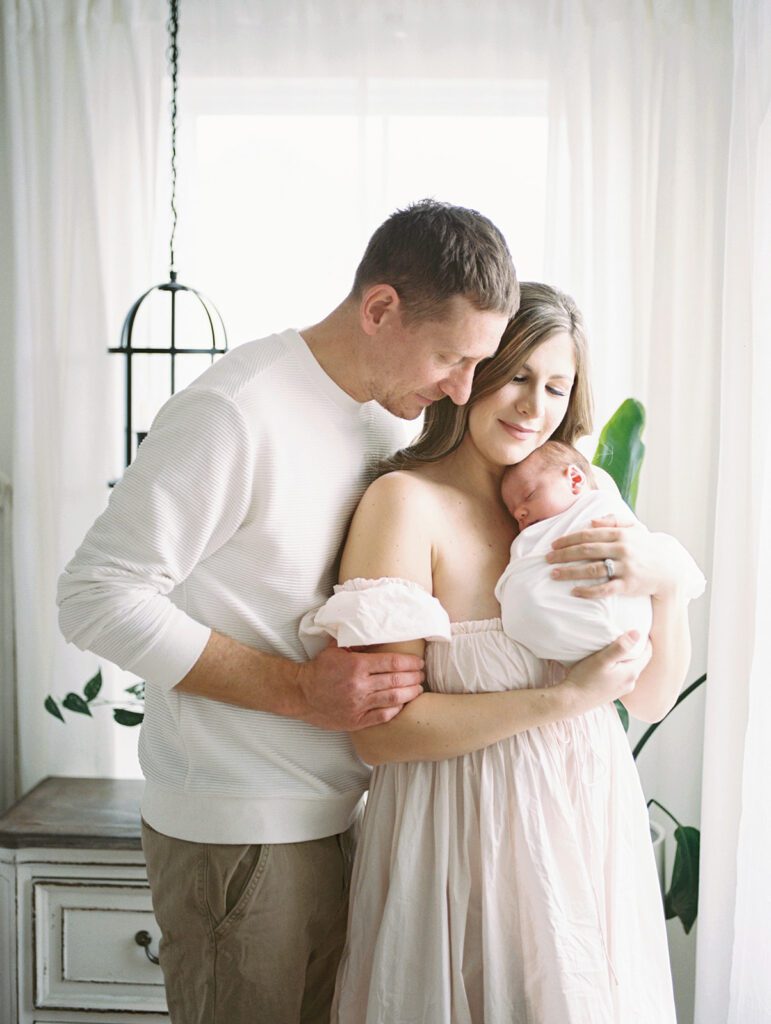 Father Stands Behind Mother, Leaning Into Her As She Holds Their Newborn Baby Girl During Their Newborn Photos In Annapolis.