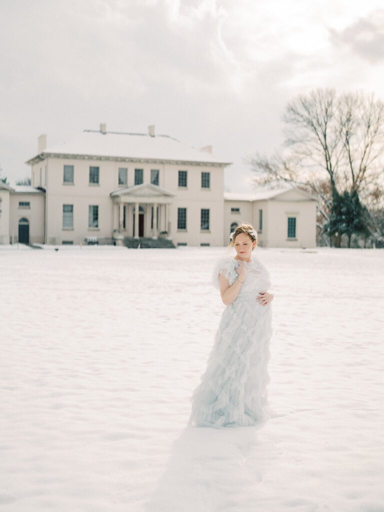 A Winter Maternity Session In The Snow Where A Mother In A Blue Needle &Amp; Thread Dress Stands In Front Of A Historic Manor.