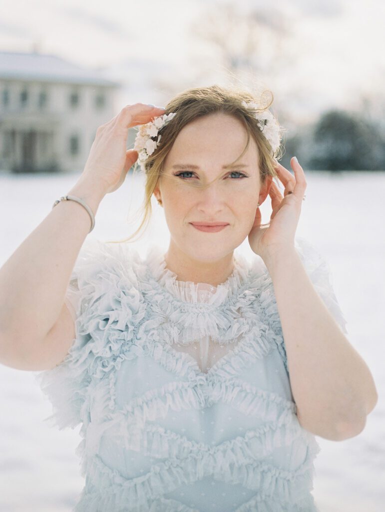 Woman With Red Hair And White Floral Crown Looks Directly At The Camera With Her Hands Adjusting Her Crown.