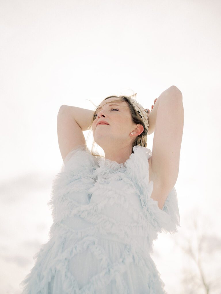 View Of Woman In Light Blue Dress Standing With Both Her Arms Behind Her Head And Her Eyes Closed.