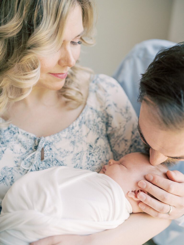 Father Leans Down To Kiss Baby's Head As Mother Holds Baby.