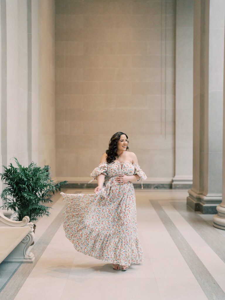 Pregnant Mother In Floral Doen Dress Walks Holding Dress And Smiling During Her National Gallery Of Art Maternity Session.