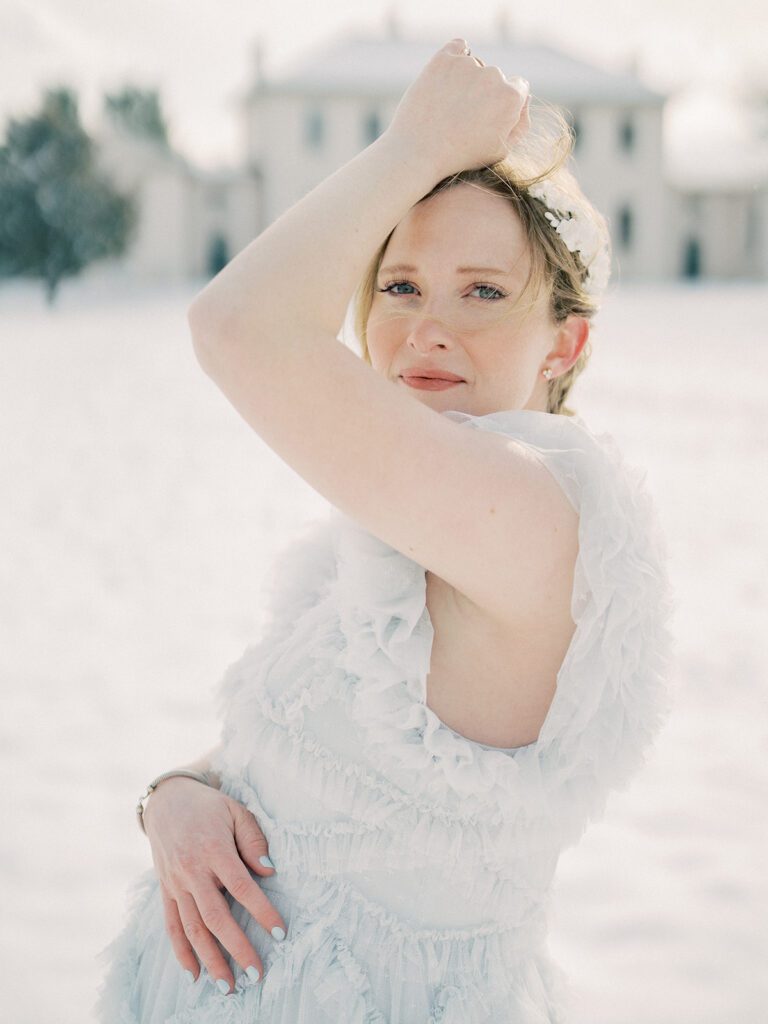 A Maryland Maternity Session With A Mom Standing In The Snow With One Hand On Top Of Her Belly And The Other Brought Up To Her Head, Looking At The Camera.