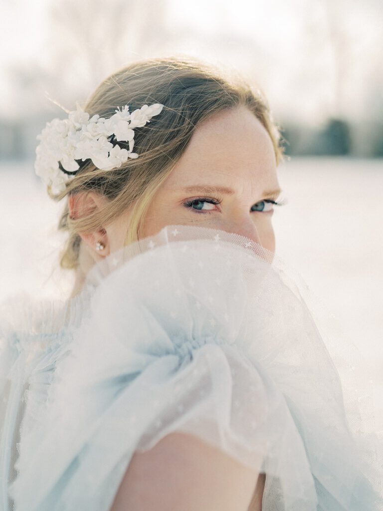 Mother With Red Hair In White Head Piece Peeks At The Camera From Behind The Shoulder Of Her Light Blue Dress During Her Winter Maternity Session In The Snow