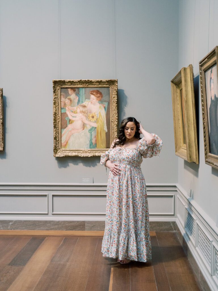 Pregnant Mother In Floral Doen Dress Stands With Hand On Belly And One Behind Her Head In The National Gallery Of Art In Dc.