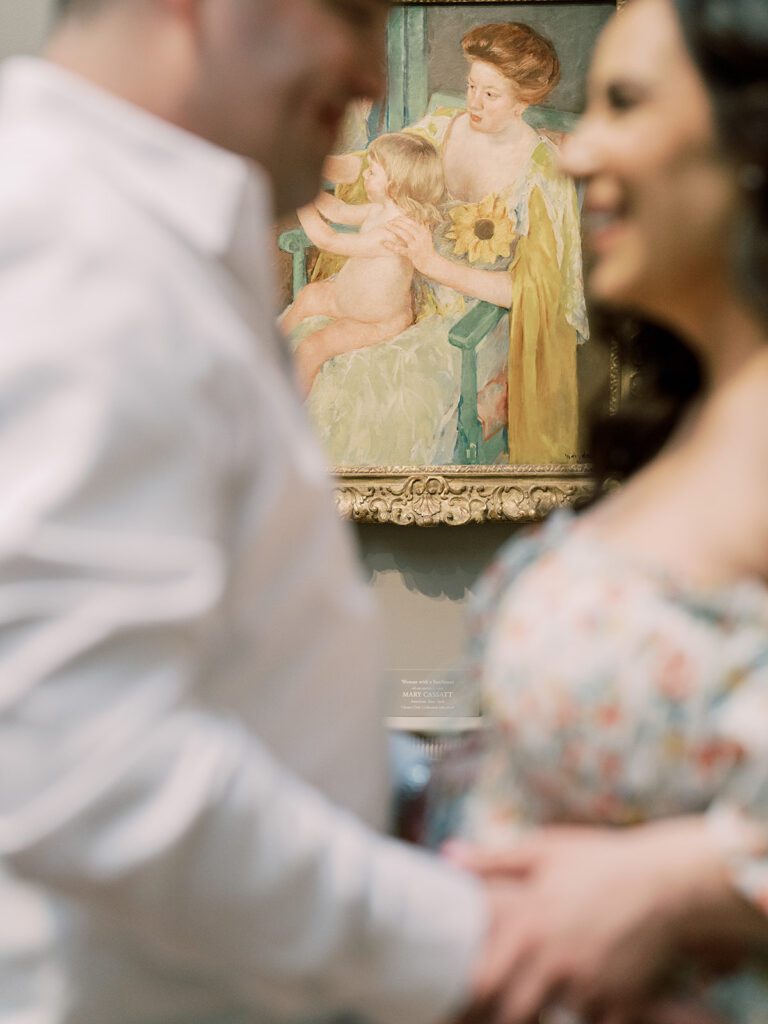 Portrait Of Mother And Child In Background Of Image While Expecting Parents Smile At Each Other In The Foreground.