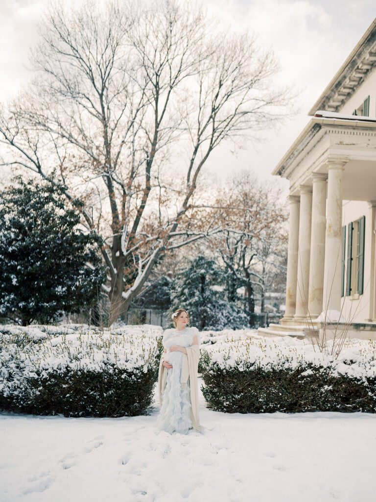 Expecting Mother Stands In Front Of Riversdale Manor In Riverdale, Maryland During Her Winter Maternity Session In The Snow.