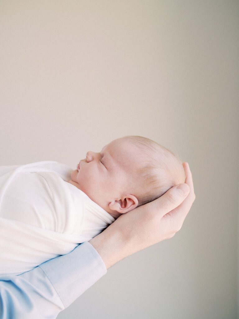 Close-Up View Of Baby Swaddled In White Held By Man In Blue Shirt.