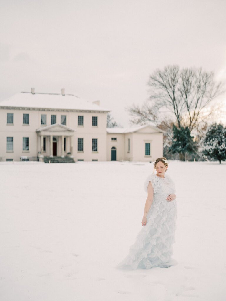 A Winter Maternity Session With A Mom Standing In Front Of Riversdale Manor In A Light Blue Dress With One Hand On Her Dress And One On Her Belly, Looking Down.