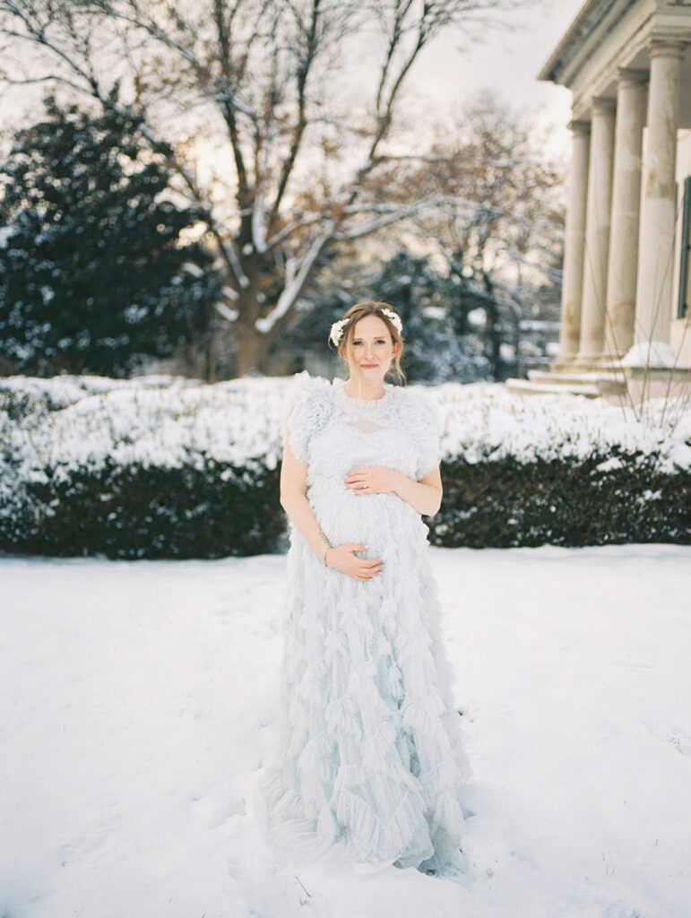 Pregnant Mother In Light Blue Dress Stands In The Snow Looking At The Camera With One Hand On Top Of Her Belly And The Other Below.