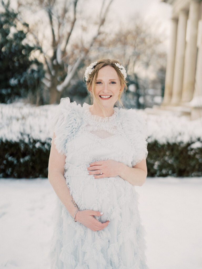 Pregnant Mother With Red Hair In Light Blue Dress And White Headpiece Smiles At The Camera With One Hand On Top Of Her Belly And The Other Below During Her Maternity Session In The Snow.