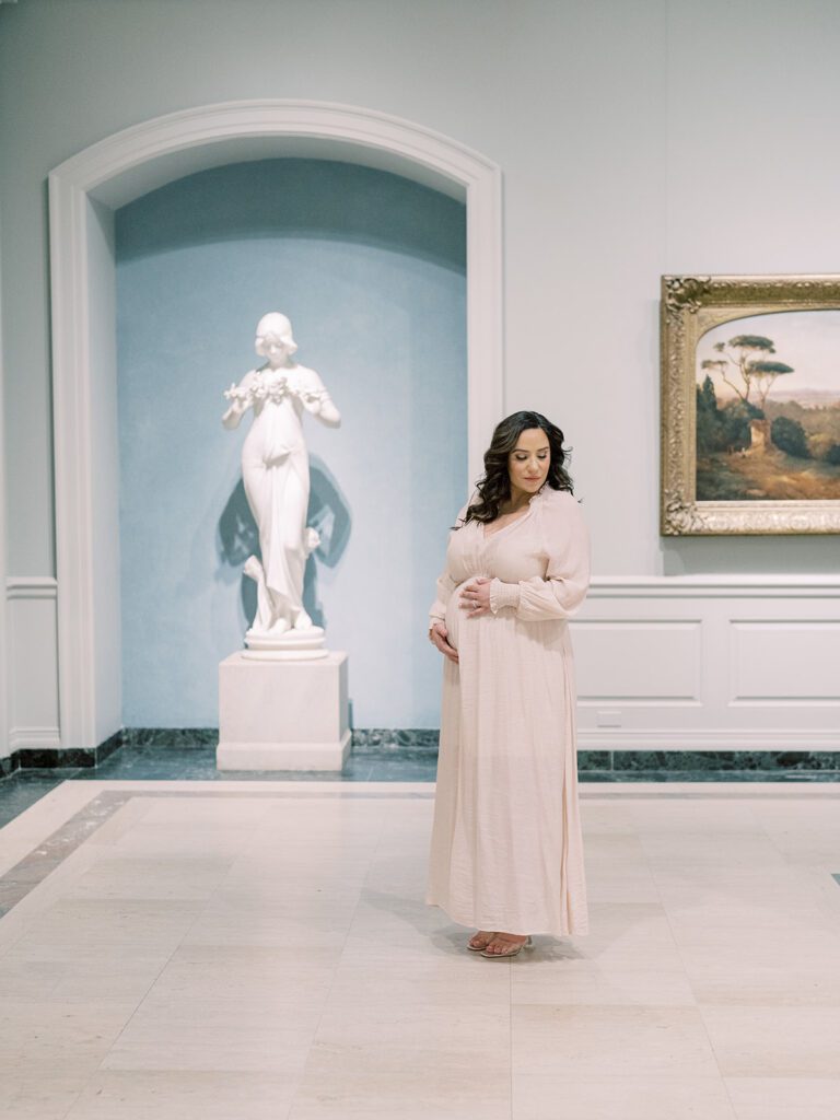 Mother With Long Brown Hair In Ivory Dress Stands In The National Gallery Of Art During Her Maternity Session.