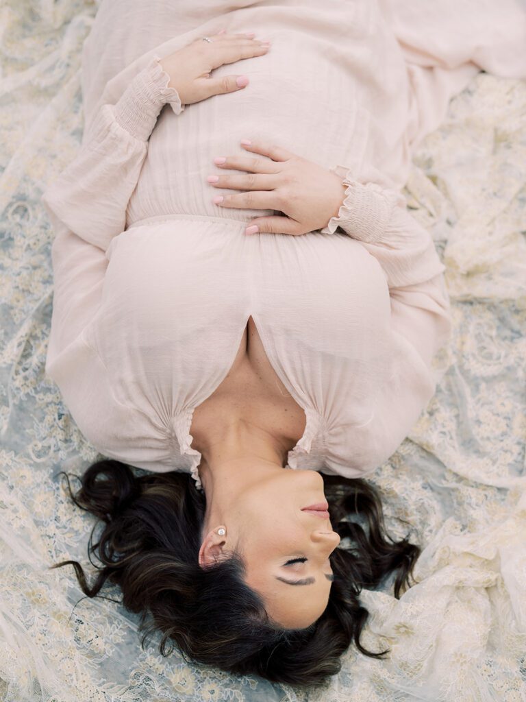 Pregnant Mother With Brown Hair Lays On Gold Lace Blanket With One Hand On Top Of Her Belly And One Below.