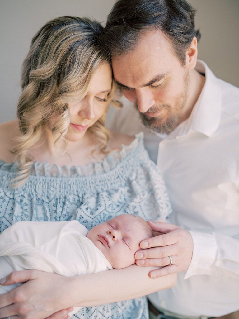 Blonde Mother And Brown-Haired Father With Beard Lean In Close And Admire Their Newborn Baby Swaddled In White.