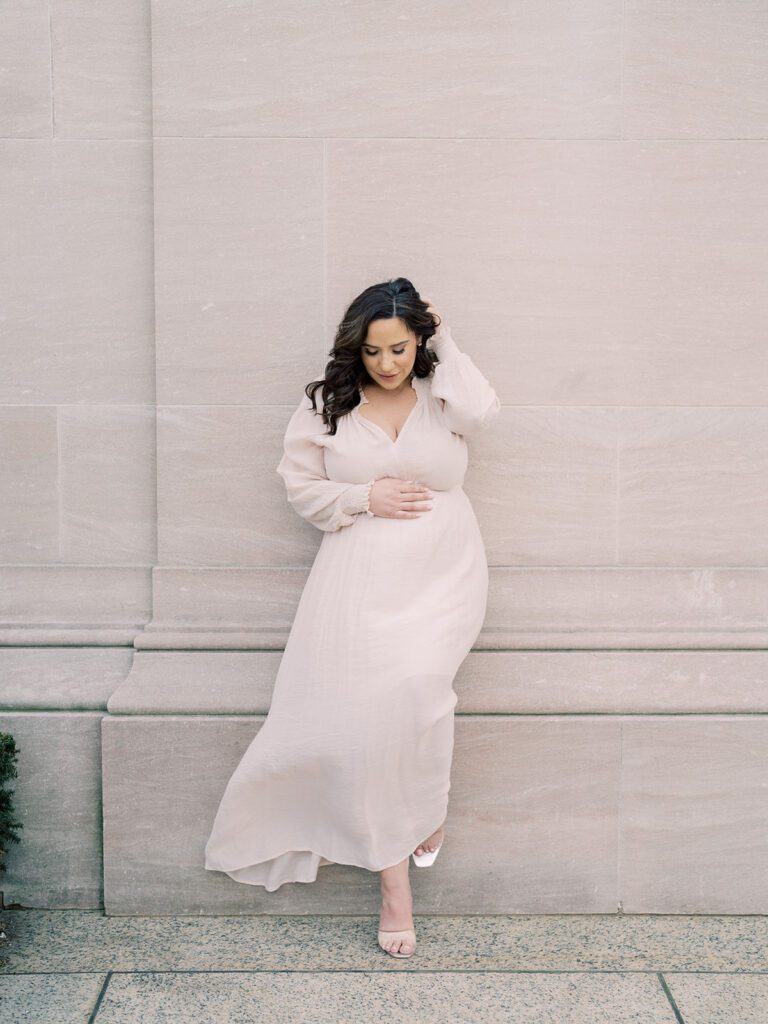 Mother With Brown Hair In Ivory Dress Leans Against Marble Wall During Her National Gallery Of Art Maternity Session.