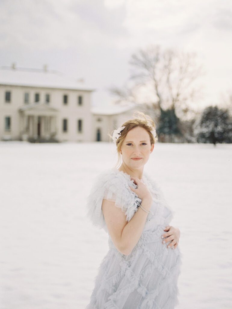 A Winter Maternity Session In The Snow With A Red-Haired Pregnant Mother Standing With One Hand On Her Belly And The Other Grazing Her Neck.