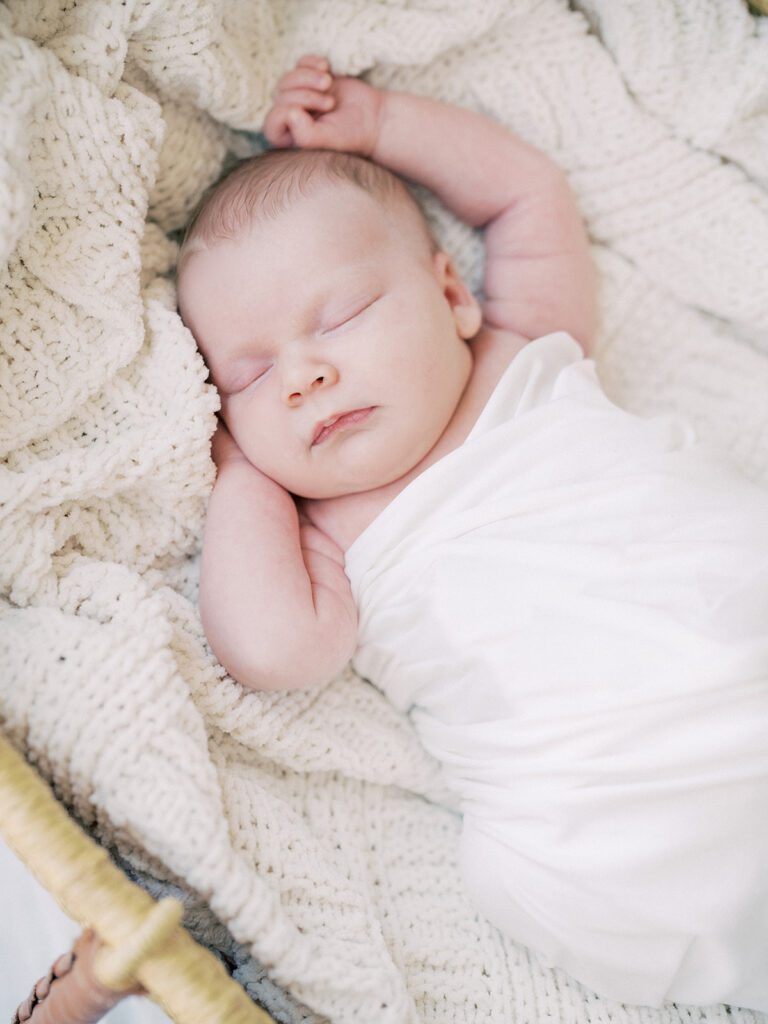 Baby Swaddled In White Sleeps With One Arm Up In A Moses Basket.