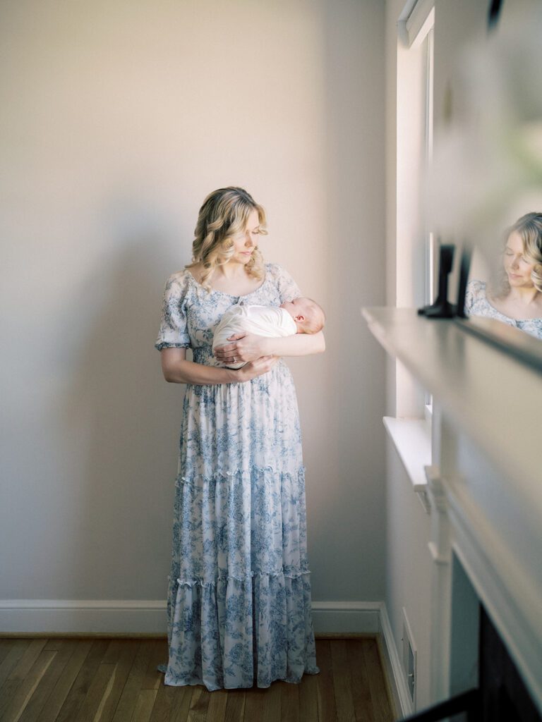 Blonde Mother Stands In Blue Floral Dress Holding Baby Swaddled In White With A View Of Her Reflection In The Mirror.