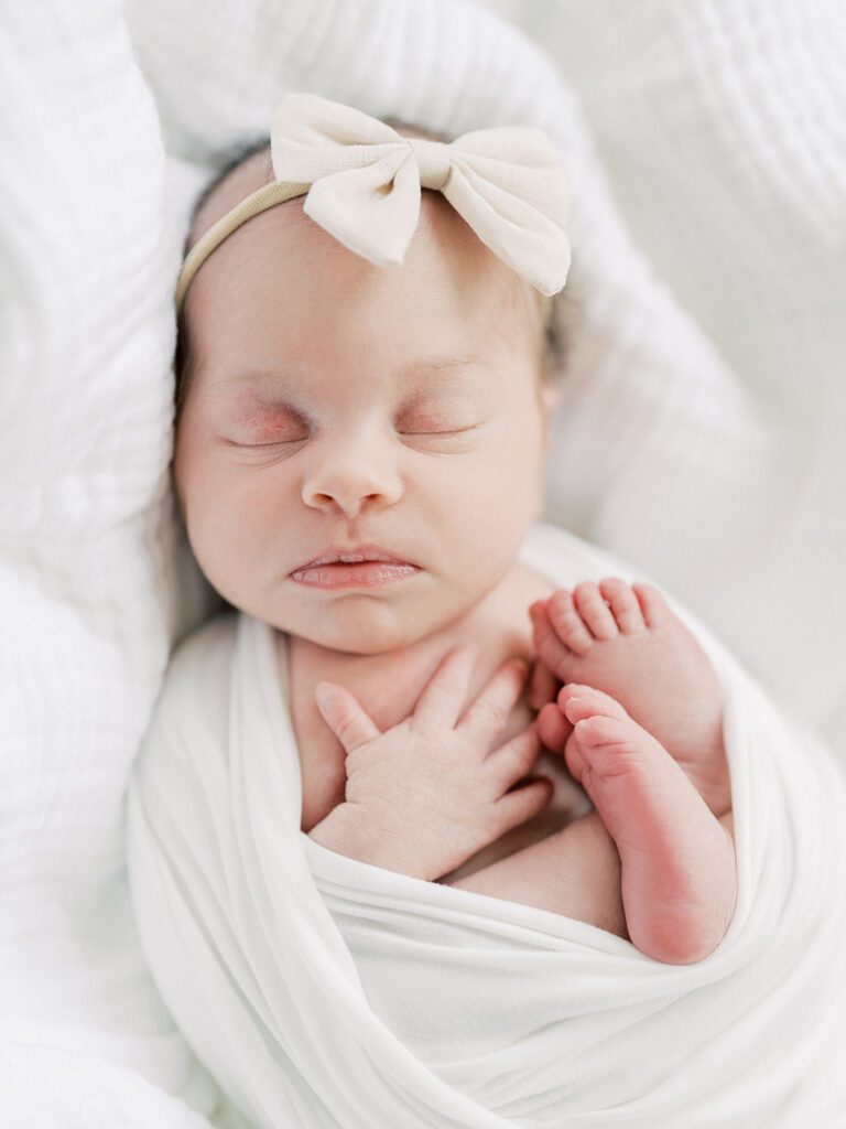 A Newborn Baby Girl Swaddled In White With A Bow On Her Head Sleeps.