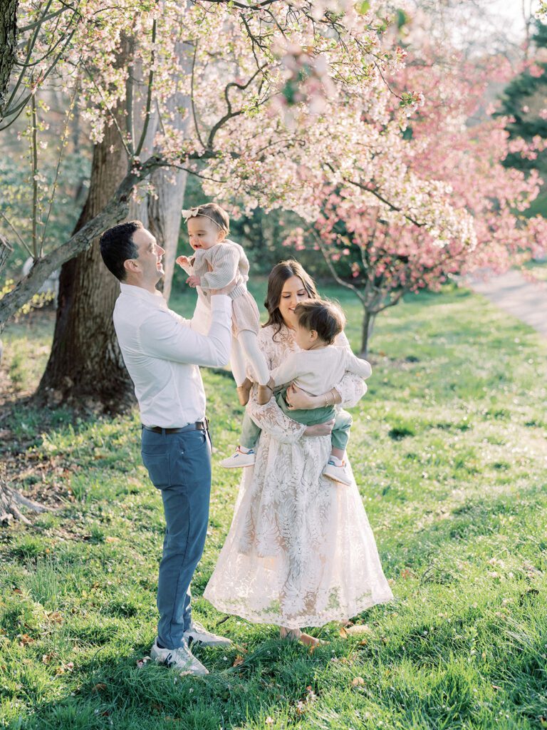 Mother And Father Dance And Swing Their Two Young Children In Front Of Cherry Blossom Trees.