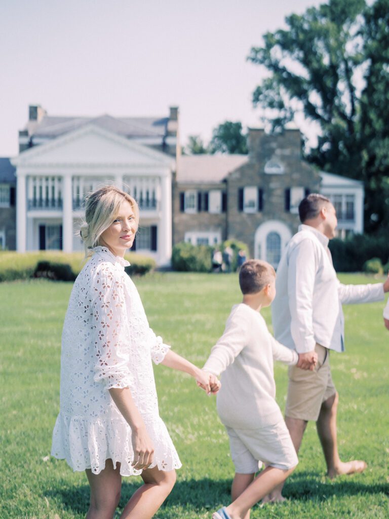 Blonde mother in white dress holds her son's hand as they walk with their family in front of Glenview Mansion MD.