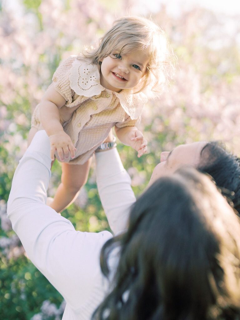 Toddler girl smiles as her parents hold her up in the air.