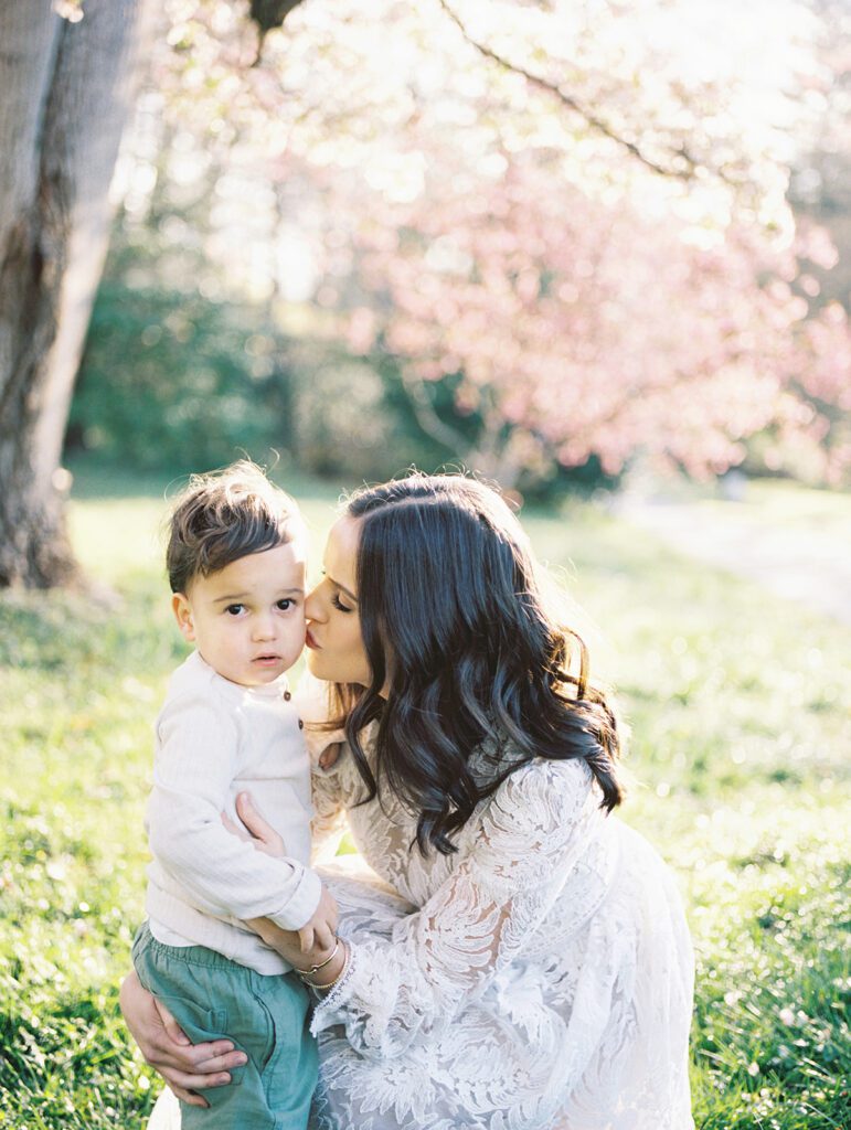 Mother With Long Brown Hair Leans In To Kiss Her Son While They Stand In Front Of Cherry Blossom Trees In Maryland At Brookside Gardens.