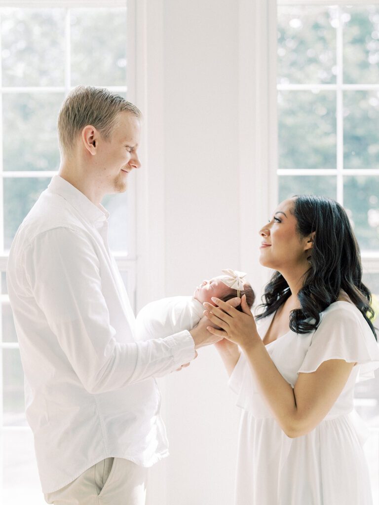 A Mother With Dark Hair Smiles Up At Her Husband As They Stand Facing Each Other Holding Their New Baby.