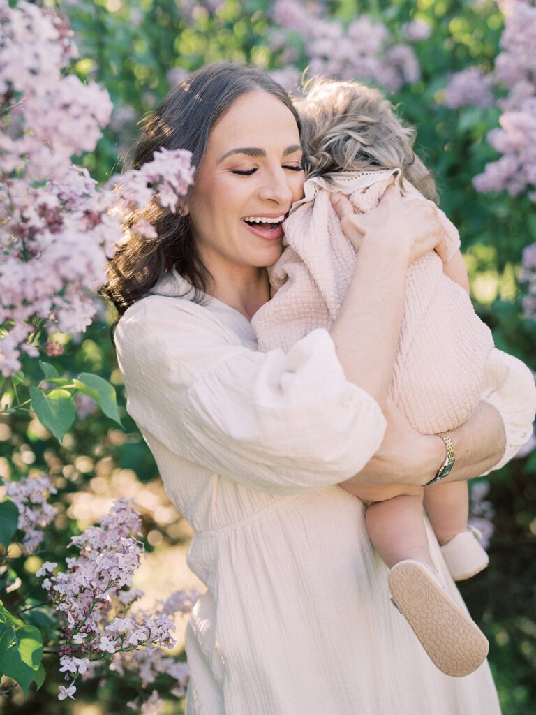 Mother Hugs Her Toddler Daughter And Smiles While Standing Near Purple Lilacs.