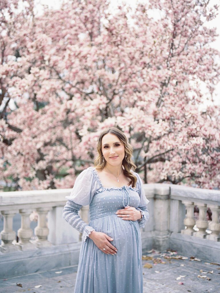 Pregnant Mother Smiles At The Camera While Wearing Blue Dress And Standing In Front Of Magnolia Trees.