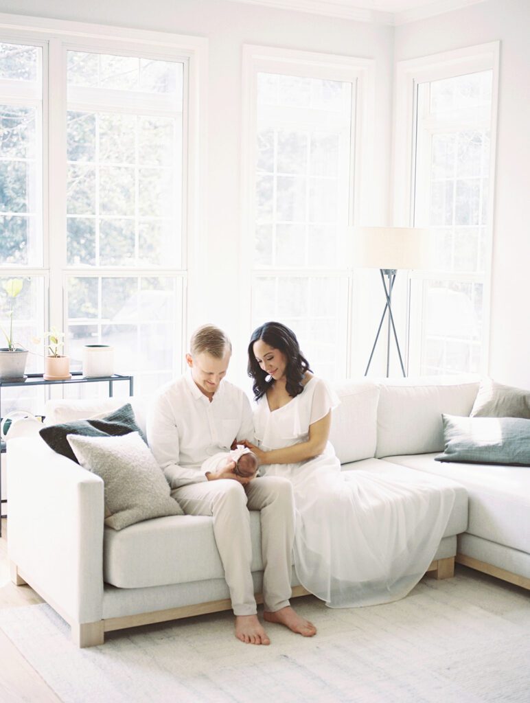 A Mother With Dark Hair In A White Dress Sits Next To Her Husband With Blonde Hair Holding Their Newborn Baby On The Couch In Their Living Room.