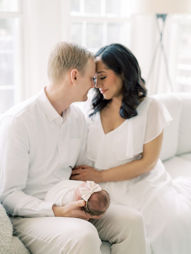 Mother And Father Lean Into Each Other While Holding Their Newborn Baby On The Couch.