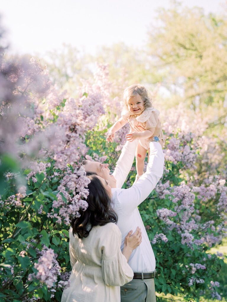 Little girl is held in the air by her parents in a field of purple lilacs, photographed by Bethesda Photographer Marie Elizabeth Photography.