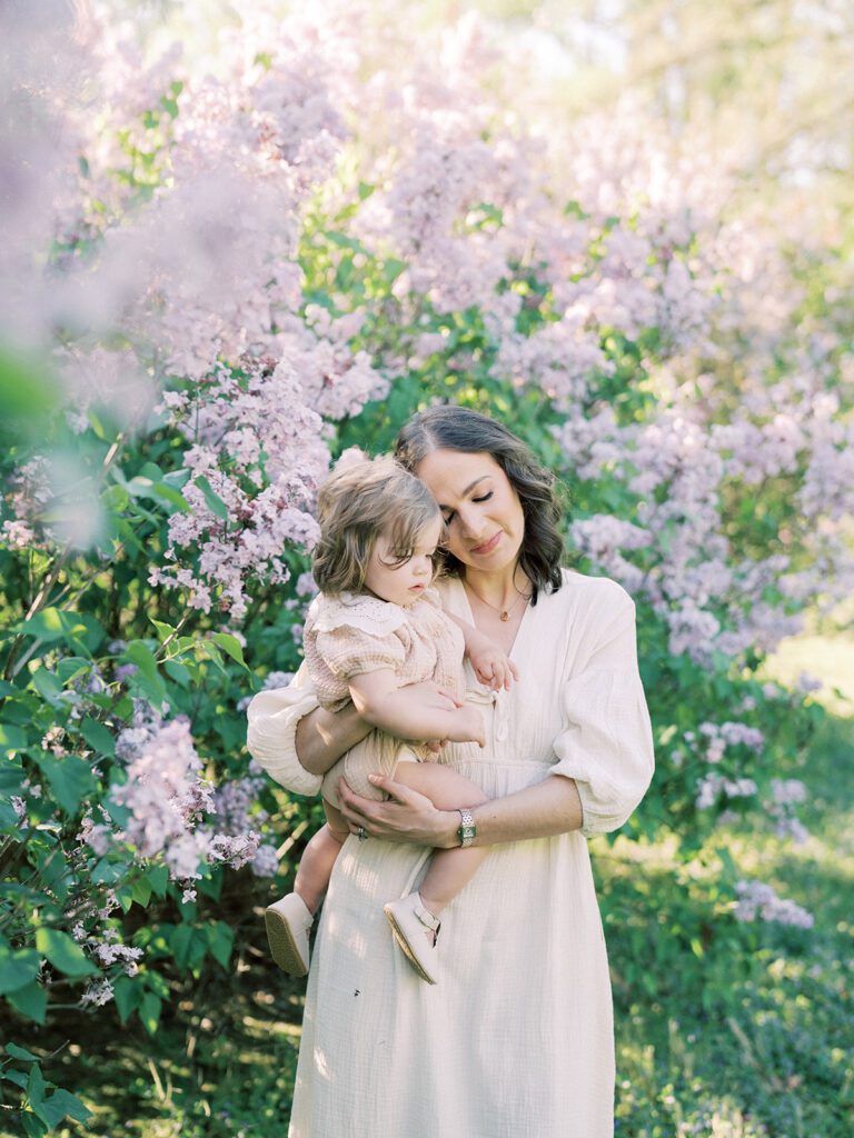 Mother With Brown Hair Leans In To Cuddle Her Toddler Daughter Who Smiles At The Camera.