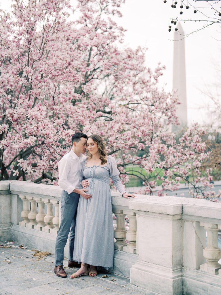 Mother In Blue Doen Dress Leans Against Stone Balcony With Her Husband At The Dar Constitution Hall With The Dc Magnolias And Washington Monument In The Background.