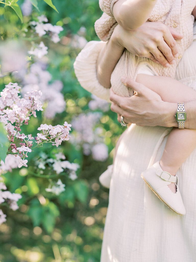 Close-Up View Of Little Girl Held By Her Mother In Front Of Purple Lilac Bush, Photographed By Bethesda Photographer Marie Elizabeth Photography.