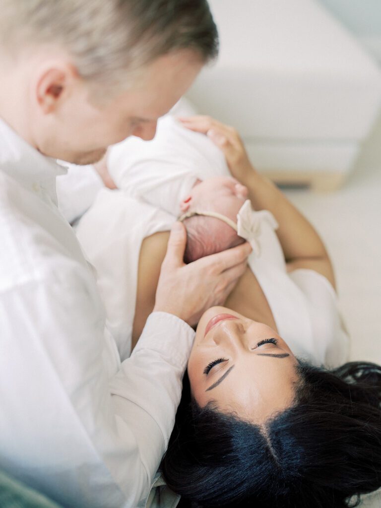 A Mother With Dark Hair Lays On Her Husband's Lap While Their Newborn Sleeps On Her Mother's Chest.